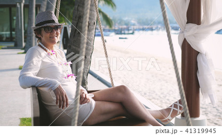 Happy woman in aged in hat and sunglasses resting on a swing on a tropical beach during summer 41319945