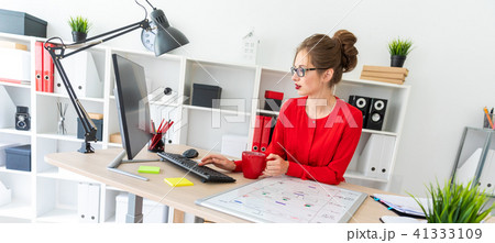 A young girl is sitting at the desk in the office, holding a red cup in her hand and typing text on A young girl is sitting at the desk in the office, holding a red cup in her hand and typing text on 41333109