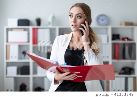 A young girl is standing in the office next to the shelving, talking on the phone and holding a 41333348