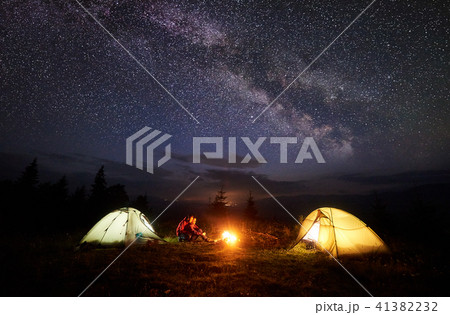 Young couple hikers resting near illuminated tent, camping in mountains at night under starry sky Young couple hikers resting near illuminated tent, camping in mountains at night under starry sky 41382232
