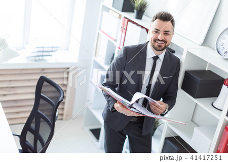 The man is standing near the rack in the office and scrolls through the folder with the documents. The man is standing near the rack in the office and scrolls through the folder with the documents. 41417251