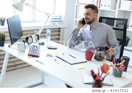 A young man in the office sits at a table, talking on the phone and holding a coffee pot. 41417332