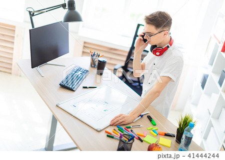 A young man in glasses stands near a computer desk and is talking on the phone. Before him lies a 41444244
