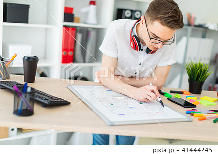 A young man in glasses stands near a computer desk. A young man draws a marker on a magnetic board 41444245
