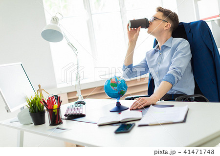 A young man sits in the office at a computer desk, holds a globe with his hand and drinks coffee. 41471748