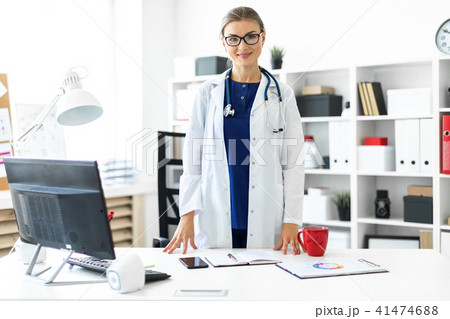 A young girl in a white coat is standing near a table in her office. A stethoscope hangs around her A young girl in a white coat is standing near a table in her office. A stethoscope hangs around her 41474688