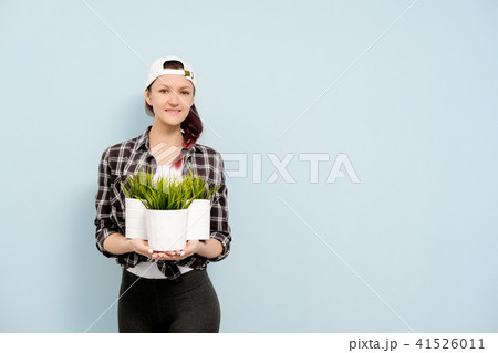 A girl in work clothes and a white cap is holding pots with green indoor plants against the blue 41526011