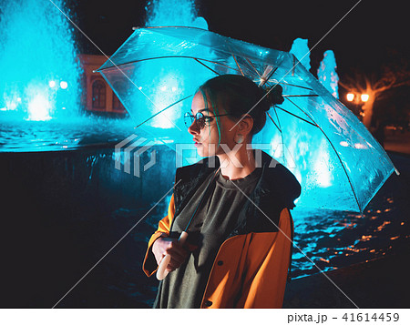 Young pretty girl with blue dyed hair in yellow raincoat and with transparent umbrella stands near 41614459