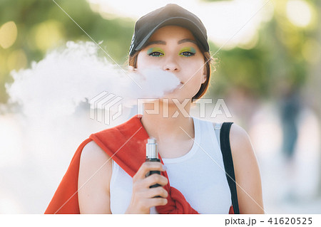 Pretty young hipster asian woman in black hat vape ecig, vaping device at the sunset. Toned image Pretty young hipster asian woman in black hat vape ecig, vaping device at the sunset. Toned image 41620525