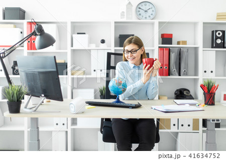 A young girl in glasses is sitting at a table, holding a red cup in her hands and looking at the 41634752