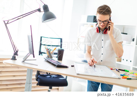 A young man in glasses stands near a computer desk and is talking on the phone. Before him lies a A young man in glasses stands near a computer desk and is talking on the phone. Before him lies a 41635689