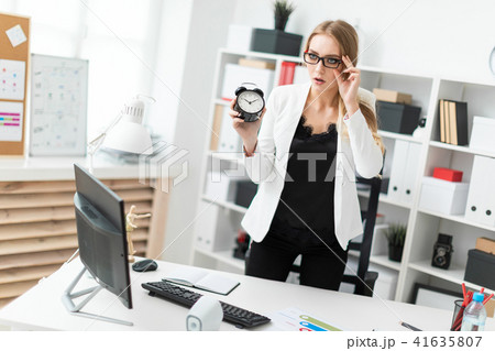 A young girl stands near a table in the office and holds an alarm clock. 41635807