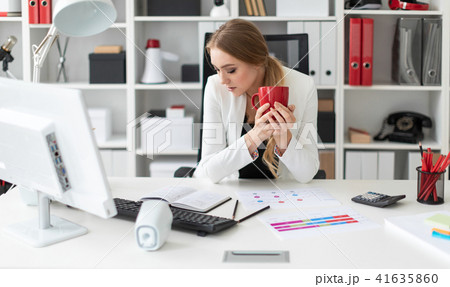 A young girl sits at a computer desk in the office and holds a red cup in her hand. Before the girl A young girl sits at a computer desk in the office and holds a red cup in her hand. Before the girl 41635860