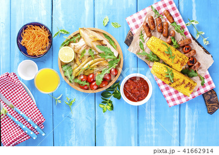 Grilled vegetables on a wooden plate and sausages, juice and salad on a blue background. Summer 41662914