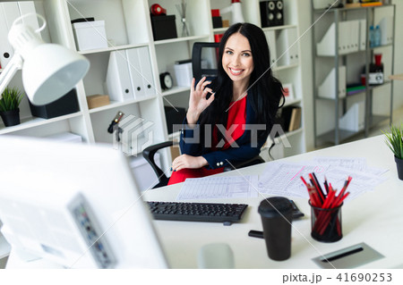 Beautiful young girl sitting in armchair at white table in office and showing okay sign. Beautiful young girl sitting in armchair at white table in office and showing okay sign. 41690253