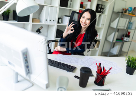 Beautiful young girl sitting in armchair at white table in office and showing okay sign. Beautiful young girl sitting in armchair at white table in office and showing okay sign. 41690255