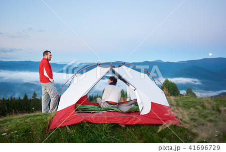 Man standing near the tent in which sits woman against beautiful scenery of mighty mountains Man standing near the tent in which sits woman against beautiful scenery of mighty mountains 41696729