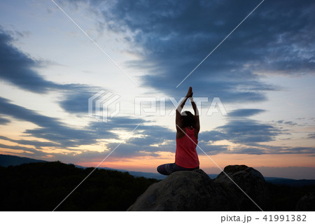 Back view of young slim athletic woman sitting on big rock in yoga pose with raised arms Back view of young slim athletic woman sitting on big rock in yoga pose with raised arms 41991382