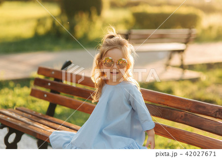 Little girl model in blue dress and sun glasses sits on a bench in the park 42027671
