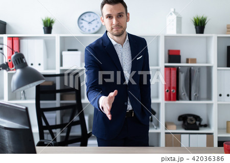 A young man stands in the office near the table and extends his hand forward. The young man says A young man stands in the office near the table and extends his hand forward. The young man says 42036386