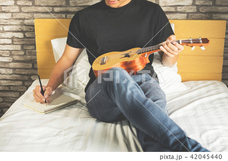 Happy young asian man playing ukulele sitting on bed in bedroom. Happy young asian man playing ukulele sitting on bed in bedroom. 42045440