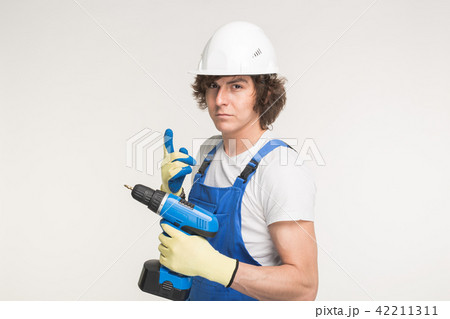 Portrait of serious builder man in white helmet with screwdriver wearing gloves and blue overalls. 42211311