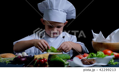 Little boy is cooking a burger at the kitchen, he is wearing chefs suit 42299213