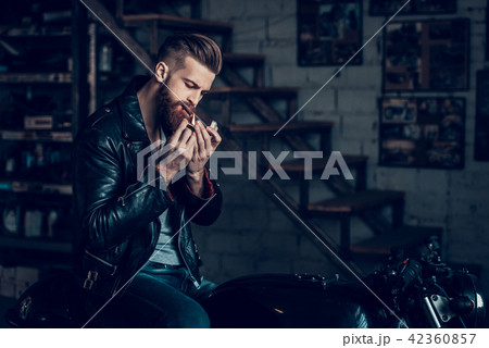 Young Bearded Biker Smoke Cigarette in Garage. Young Bearded Biker Smoke Cigarette in Garage. 42360857