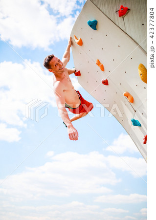 Photo of smiling athlete looking at camera practicing on wall for rock climbing against blue sky 42377804