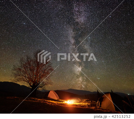 Night camping in the mountains. Hikers having a rest in tourist tent under starry sky near campfire Night camping in the mountains. Hikers having a rest in tourist tent under starry sky near campfire 42513252