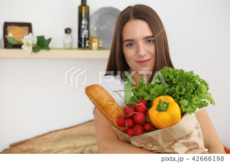 Young woman cooking in kitchen. Girl holding paper bag full of vegetables and fruits. Householding Young woman cooking in kitchen. Girl holding paper bag full of vegetables and fruits. Householding 42666198