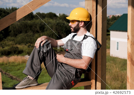 The man builder sits on the edge of the roof of the frame house, in a yellow helmet and gray The man builder sits on the edge of the roof of the frame house, in a yellow helmet and gray 42692573