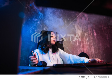 Young brunette woman wearing hoodie, sitting down by desk with computer while smoking e-cigarette Young brunette woman wearing hoodie, sitting down by desk with computer while smoking e-cigarette 42700661