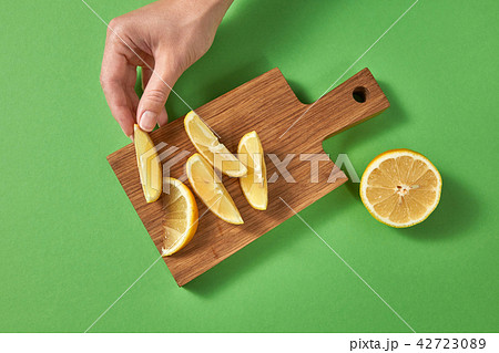 Slices of lemon on a board on a green table. A female hand puts slice of ripe fresh lemon on a 42723089