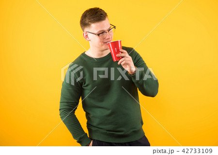Portrait of young handsome confident guy in green sweater holding red plastic cup on yellow 42733109