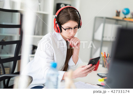 A young girl sits in headphones at a table in the office and holds a phone in her hand. 42733174