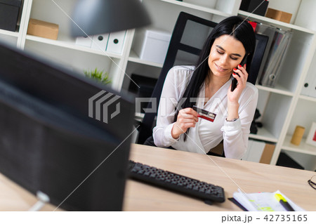 A young girl is sitting at a table, holding a bank card in her hand and talking on the phone. 42735165
