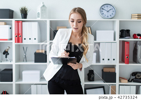 A young girl stands in the office next to the shelf and holds documents and a pen in her hand. A young girl stands in the office next to the shelf and holds documents and a pen in her hand. 42735324