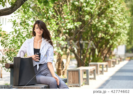 Pretty brunette drinking coffee sitting on wooden bench near paper bags. 42774602