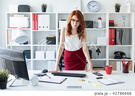 A beautiful young girl stands in the office near the table, putting her hands on it. A beautiful young girl stands in the office near the table, putting her hands on it. 42786666