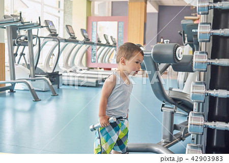 Little child standing in gym in front of metallic dumbbells and racetracks. Little child standing in gym in front of metallic dumbbells and racetracks. 42903983