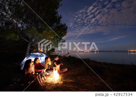 Night summer camping on shore. Group of young tourists around campfire near tent under evening sky Night summer camping on shore. Group of young tourists around campfire near tent under evening sky 42917534