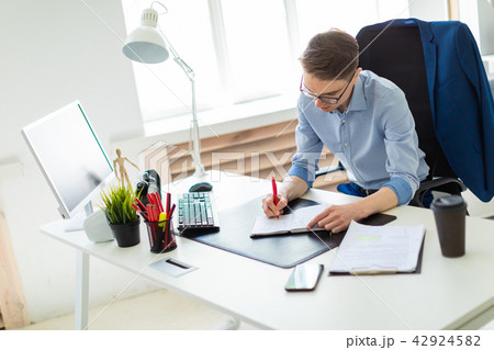 A young man sits in the office at a computer desk and writes in a notebook. A young man sits in the office at a computer desk and writes in a notebook. 42924582
