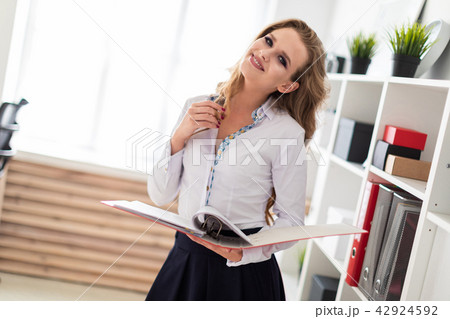 A beautiful young girl stands near a rack in the office and holds a folder with documents. 42924592