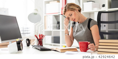 A young girl sits at a table in the office and holds a red cup and glasses in her hands. Before the A young girl sits at a table in the office and holds a red cup and glasses in her hands. Before the 42924642