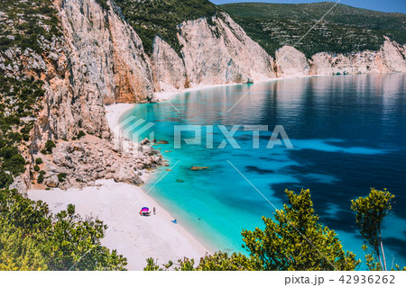 Amazing Fteri beach lagoon, Kefalonia, Greece. Tourists under umbrella chill relax near clear blue Amazing Fteri beach lagoon, Kefalonia, Greece. Tourists under umbrella chill relax near clear blue 42936262