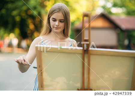 Beautiful girl draws a picture in the park using a palette with paints and a spatula. Easel and 42947339