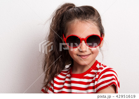A small girl with red sunglasses and striped T-shirt standing in a studio. 42951619