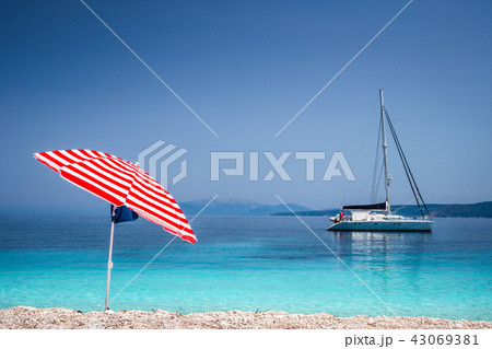 Bright red striped sun beach umbrella on pebble beach against turquoise blue shellow sea water and Bright red striped sun beach umbrella on pebble beach against turquoise blue shellow sea water and 43069381