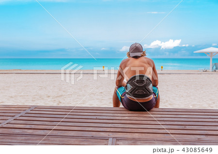 The surfer sits with his back to the camera on the terrace on the beach looking out into the The surfer sits with his back to the camera on the terrace on the beach looking out into the 43085649
